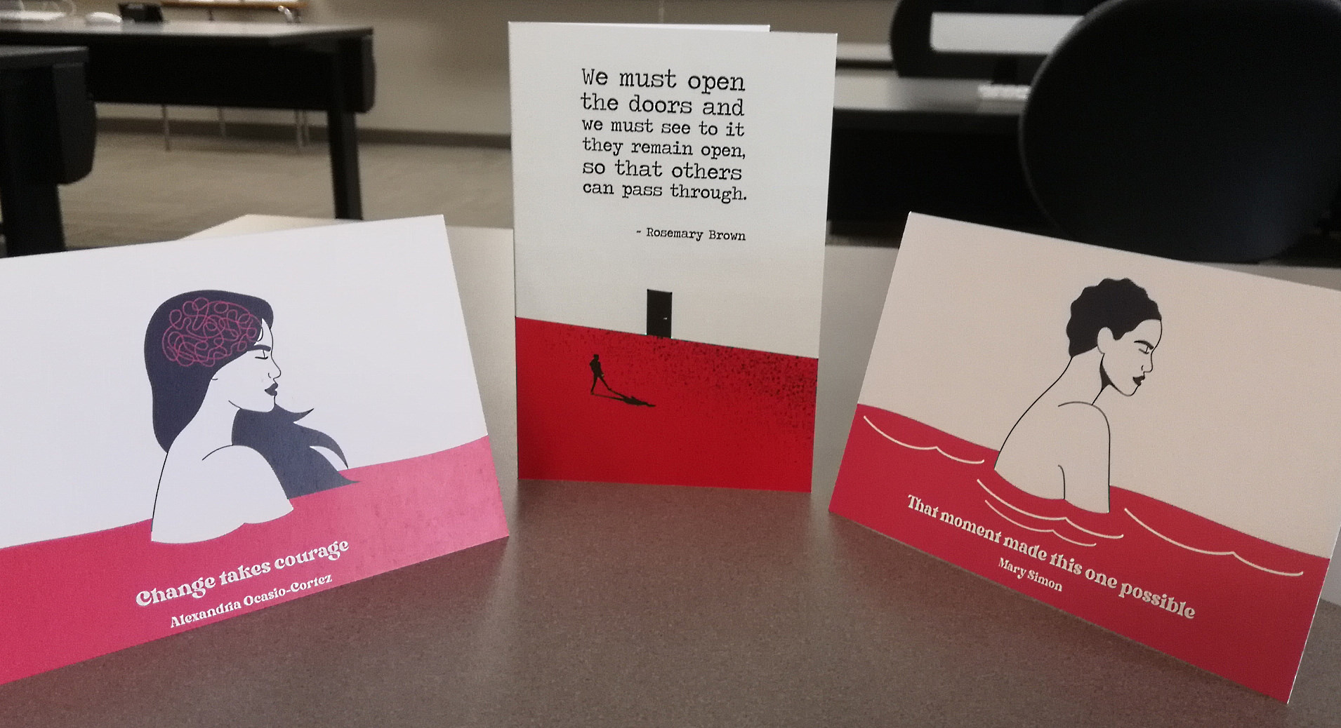 Three greeting cards sitting upright on a desk. Each card has quotes from famous women with stark shape-based illustrations of Women moving through doors, climbing mountains, and sitting alone.